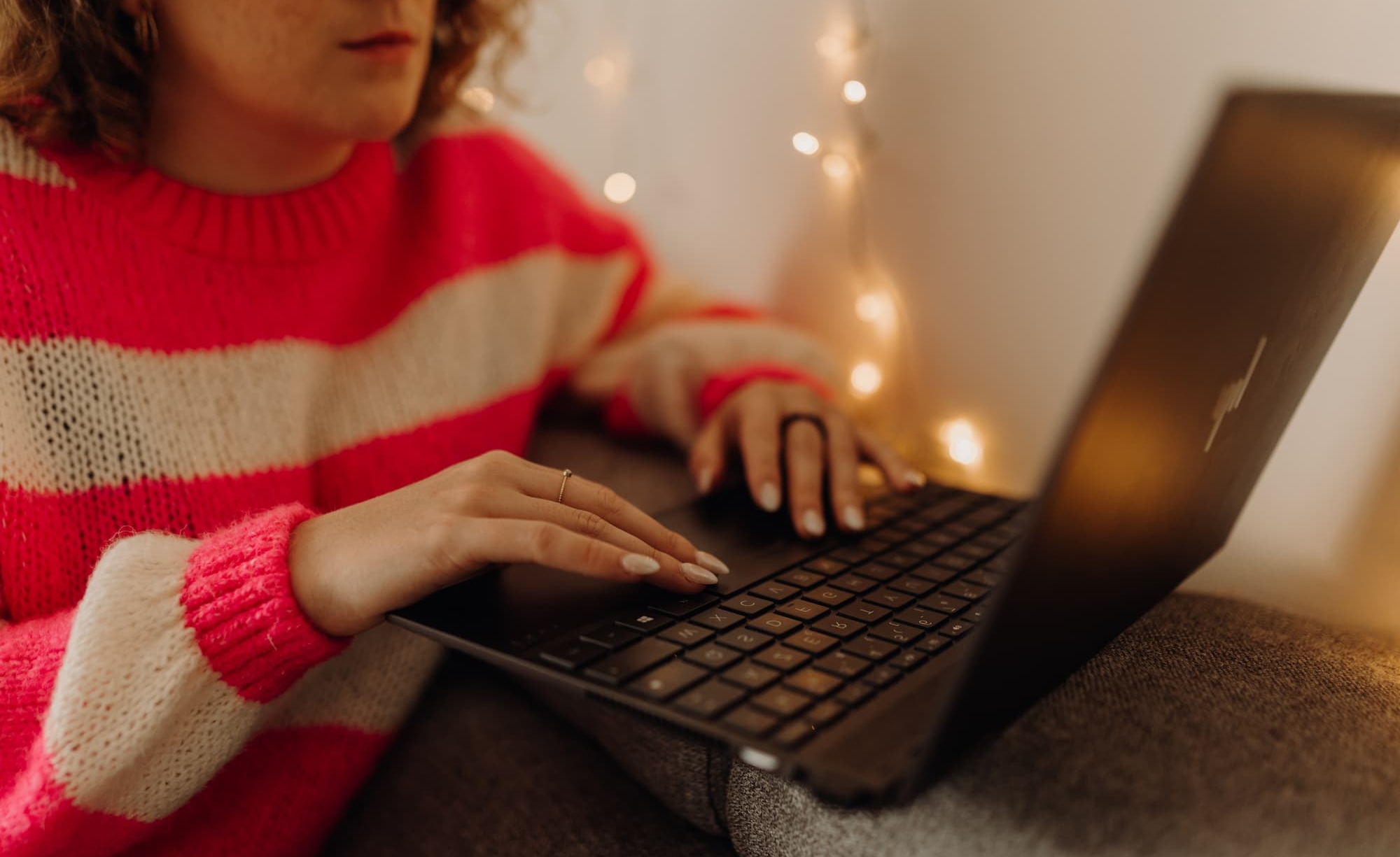 Gros plan sur les mains de Clothilde, en pull rayé rose, tapant sur le clavier de son ordinateur portable devant une guirlande lumineuse