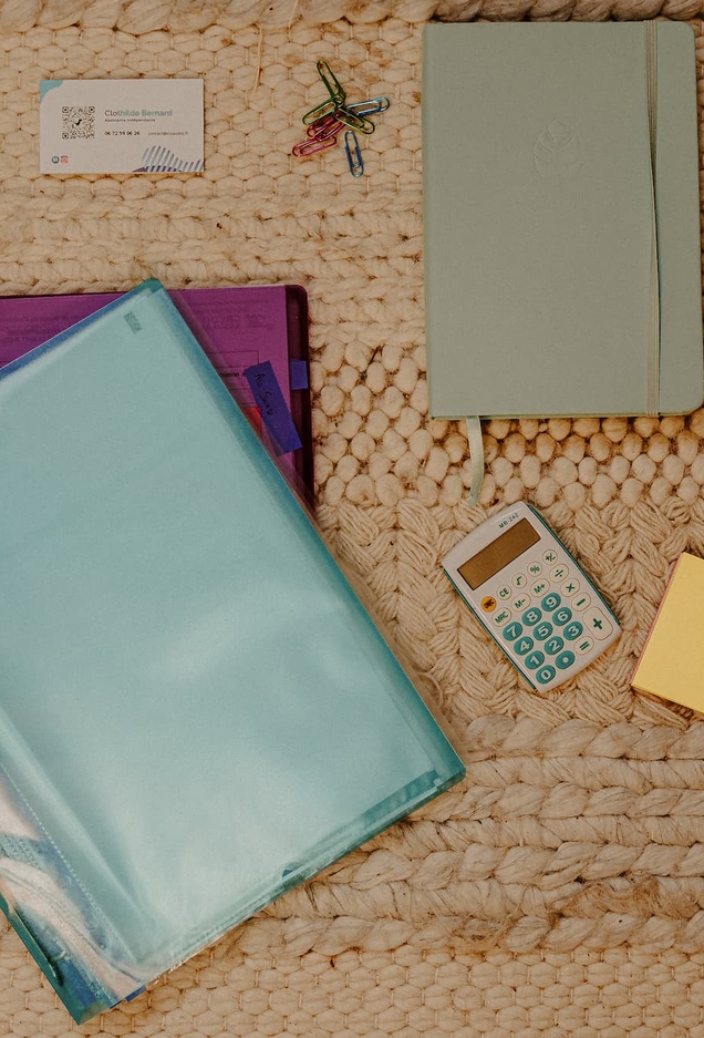 Fournitures d'une assistante administrative posées sur un tapis tressé : carte de visite CloAssist', calculatrice, carnet, pochettes et trombones