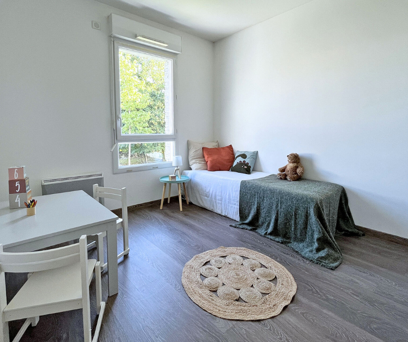 Chambre d’enfant décorée avec sobriété grâce au Home staging à Nantes  : lit simple avec jeté vert et coussins colorés, tapis en jute rond, table enfant blanche et vue sur verdure.