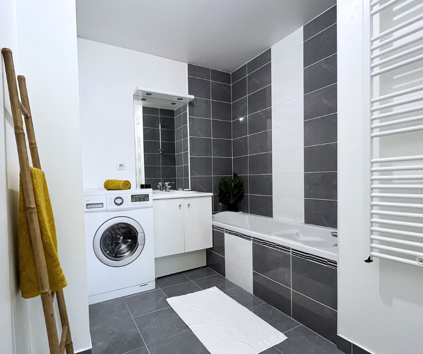 Salle de bains moderne mise en valeur après home staging à Nantes avec baignoire, carrelage gris et blanc, meuble vasque blanc, lave-linge en carton imprimé, porte-serviette mural et échelle en bambou décorative.
