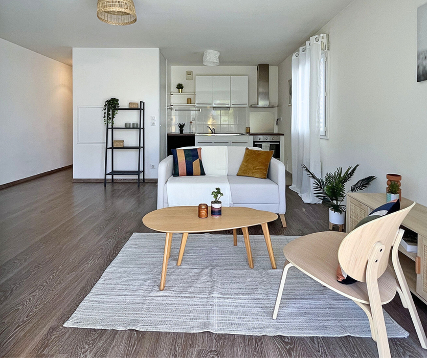 Séjour lumineux mis en scène à Nantes : canapé beige et tables basses sur un tapis clair, avec meuble TV et étagère, planchers foncés, murs blancs et grandes baies vitrées laissant entrer la lumière naturelle
