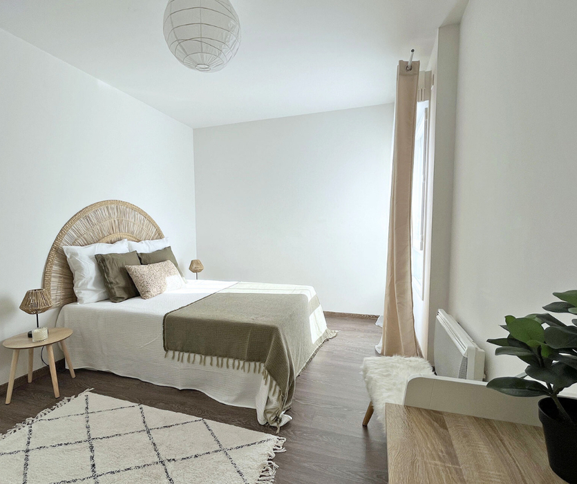 Chambre parentale lumineuse et épurée après home staging à Nantes : lit double avec tête de lit en rotin, linge de lit aux tons naturels, chevets en bois, rideaux beiges et parquet foncé.
